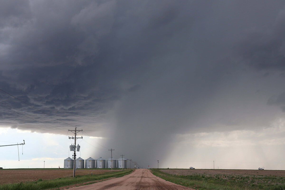 A dark cloud system dumping rain in a slanting column onto the horizon of farmland with six grain silos in a line.